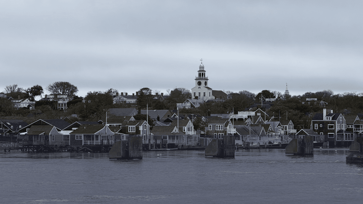 Gray harbor under a cloudy sky, with shingled waterfront houses and a white church clock tower on the hill.